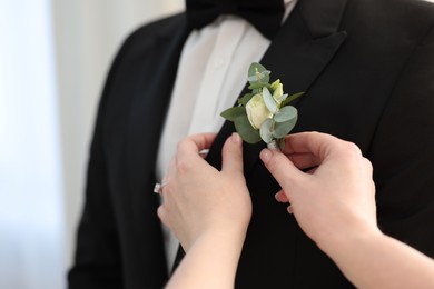Bride putting boutonniere on her groom's jacket against light background, closeup. Wedding accessory