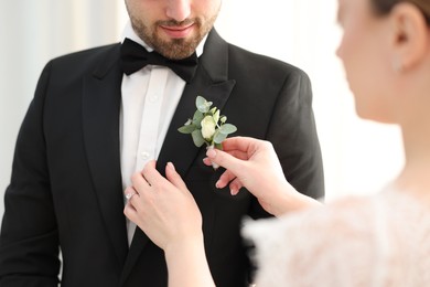 Photo of Bride putting boutonniere on her groom's jacket against light background, closeup. Wedding accessory