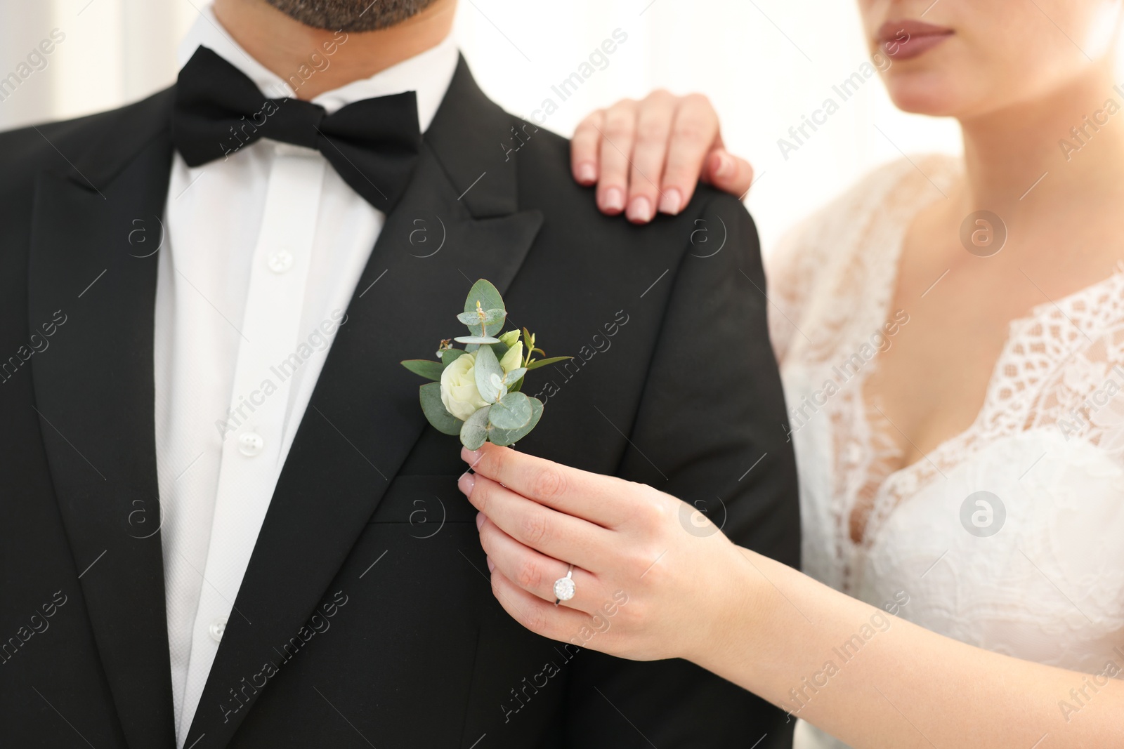 Photo of Bride putting boutonniere on her groom's jacket against light background, closeup. Wedding accessory