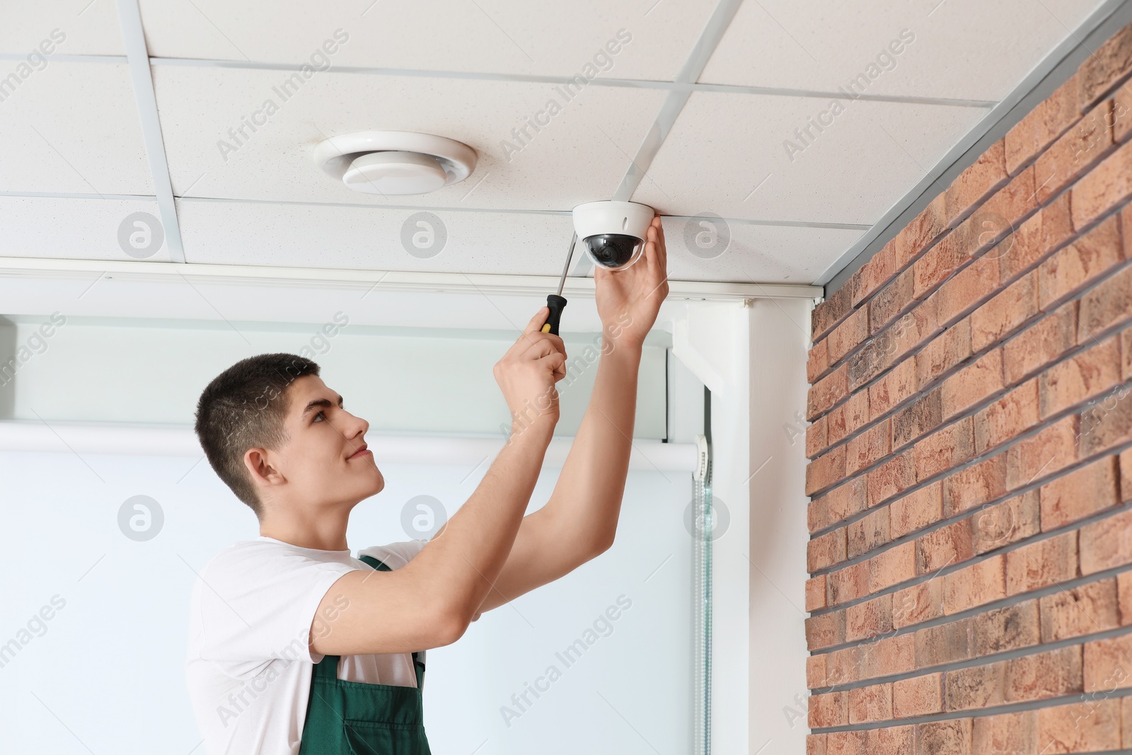 Photo of Technician with screwdriver installing CCTV camera on ceiling indoors