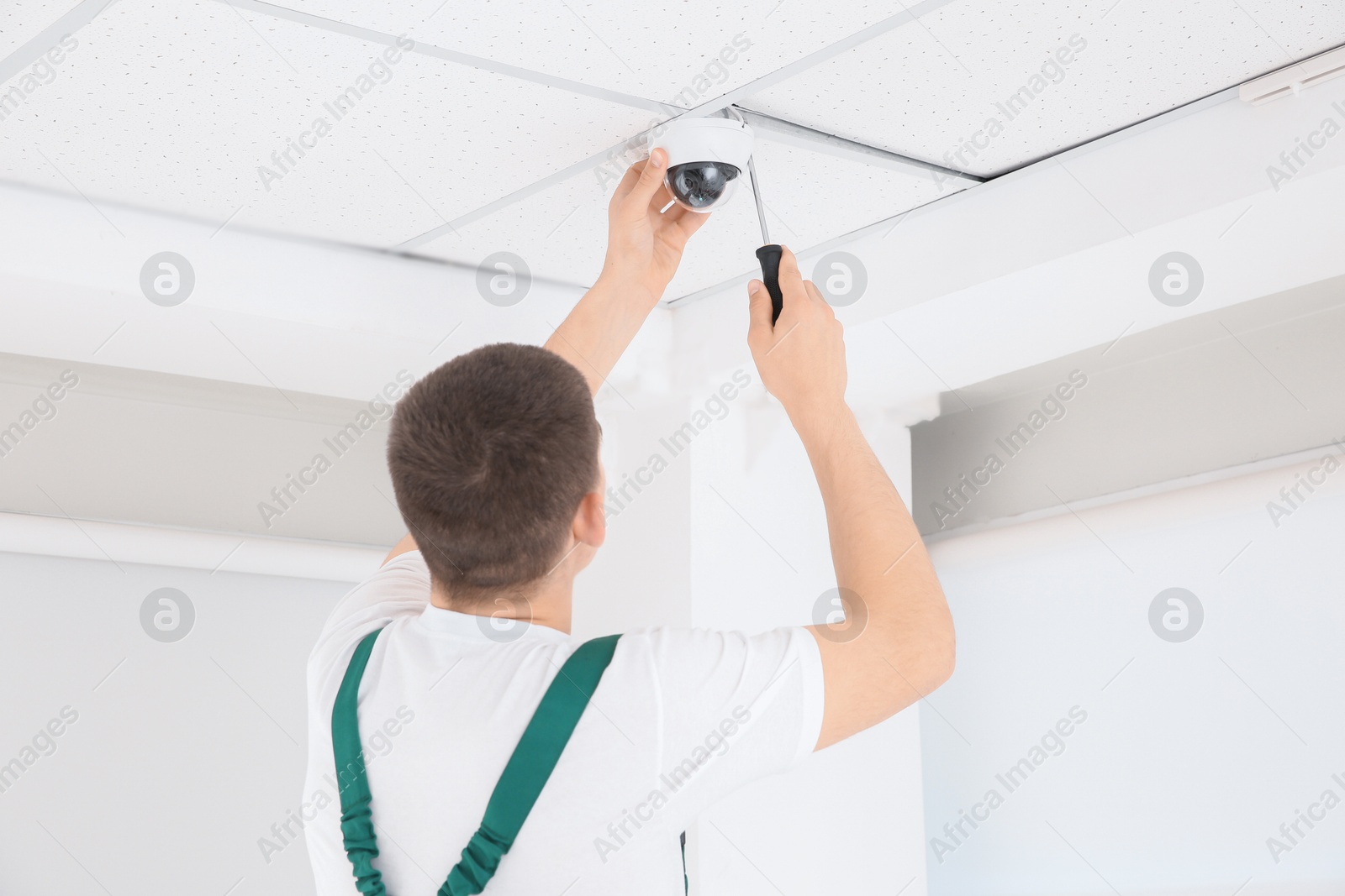 Photo of Technician with screwdriver installing CCTV camera on ceiling indoors, back view