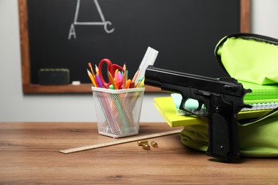 Photo of Gun, bullets and school stationery on wooden table near blackboard indoors. Space for text