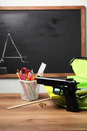 Photo of Gun, bullets and school stationery on wooden table near blackboard indoors