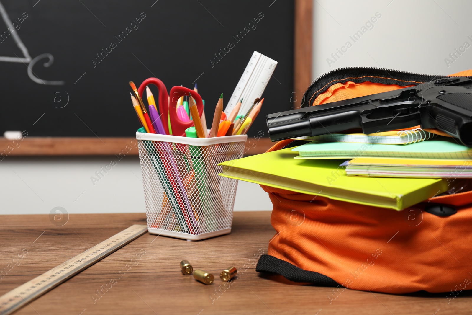 Photo of Gun, bullets and school stationery on wooden table near blackboard indoors, closeup