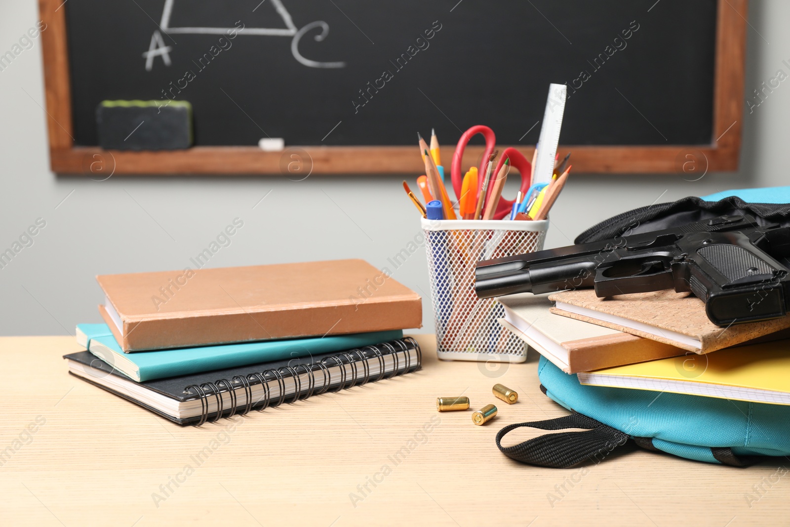 Photo of Gun, bullets and school stationery on wooden table near blackboard indoors