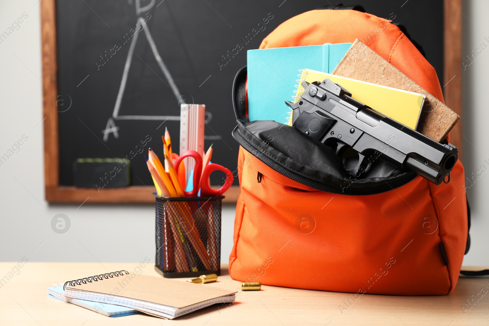 Photo of Gun, bullets and school stationery on wooden table near blackboard indoors