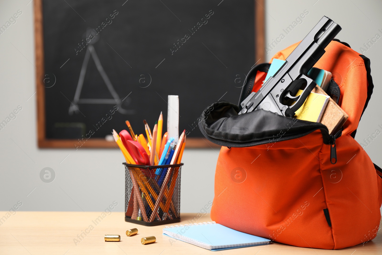 Photo of Gun, bullets and school stationery on wooden table near blackboard indoors, space for text