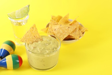 Photo of Delicious guacamole with nachos chips, maracas, tequila and lime on yellow background, space for text