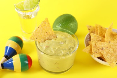 Delicious guacamole with nachos chips, maracas, tequila and lime on yellow background,