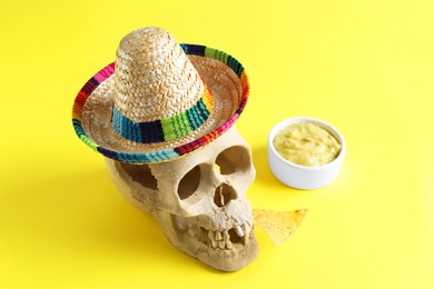Photo of Human scull with Mexican sombrero hat, nachos chip and guacamole on yellow background