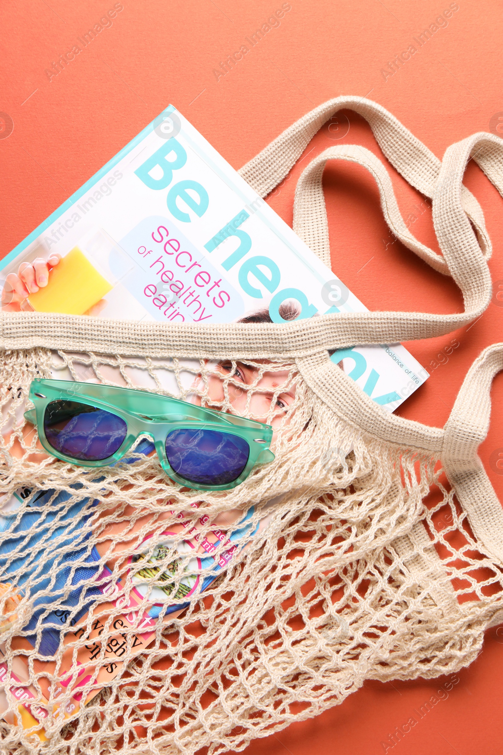 Photo of String bag, sunglasses and magazine on coral background, top view