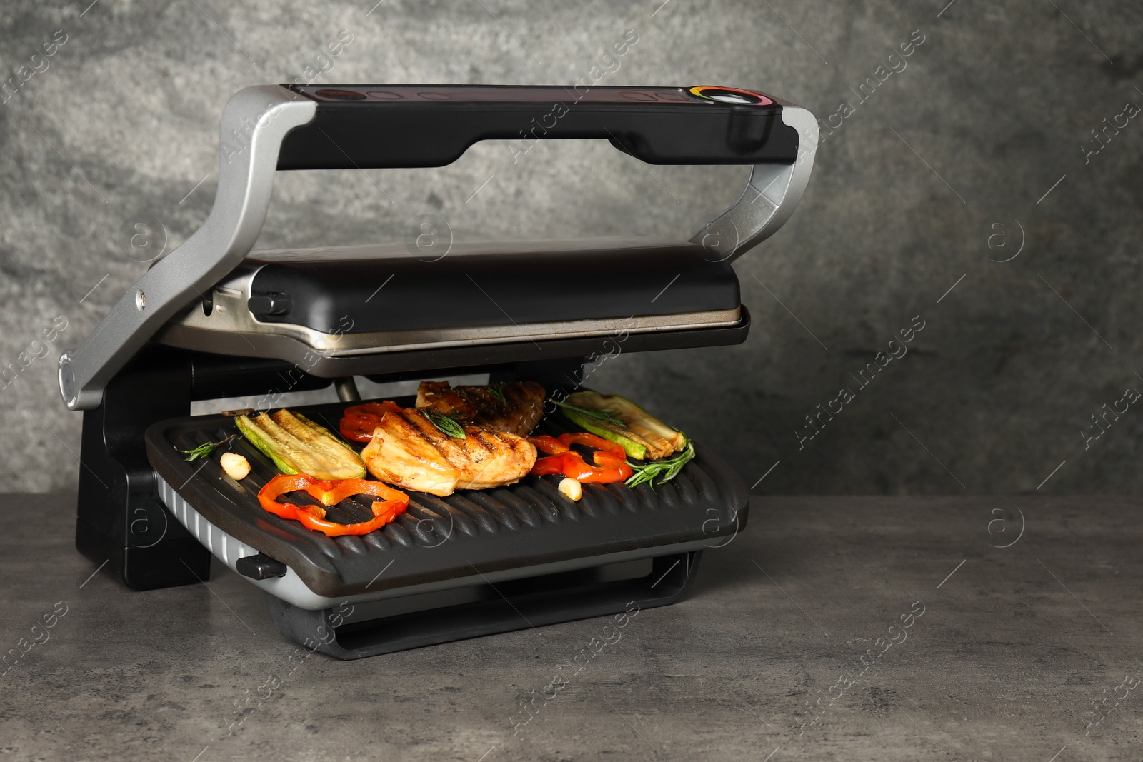 Photo of Electric grill with tasty meat, spices and vegetables on grey textured table, space for text
