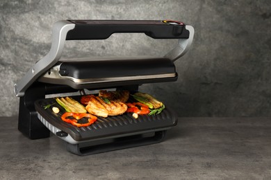 Photo of Electric grill with tasty meat, spices and vegetables on grey textured table, space for text