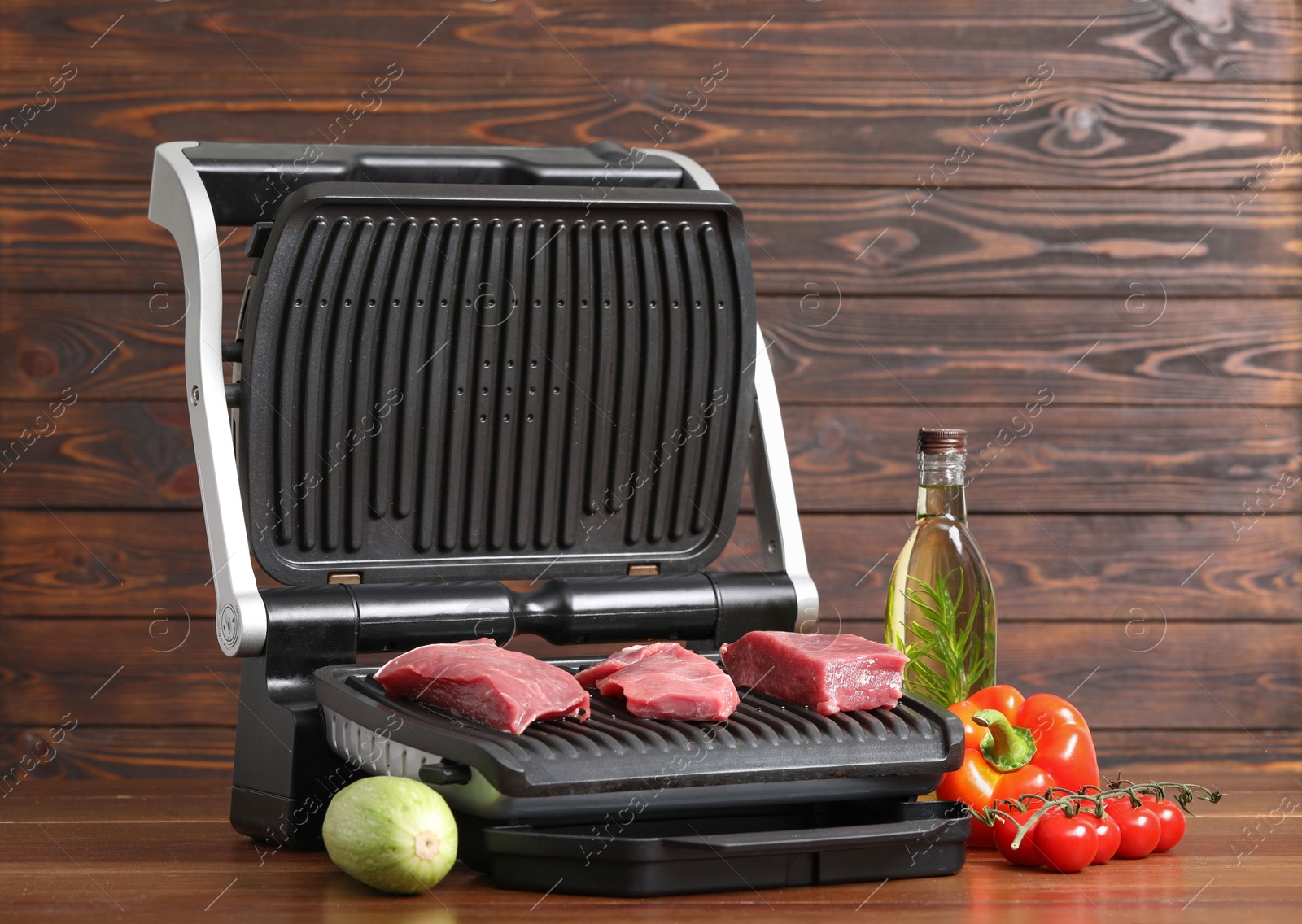 Photo of Electric grill with raw meat, oil and vegetables on wooden table