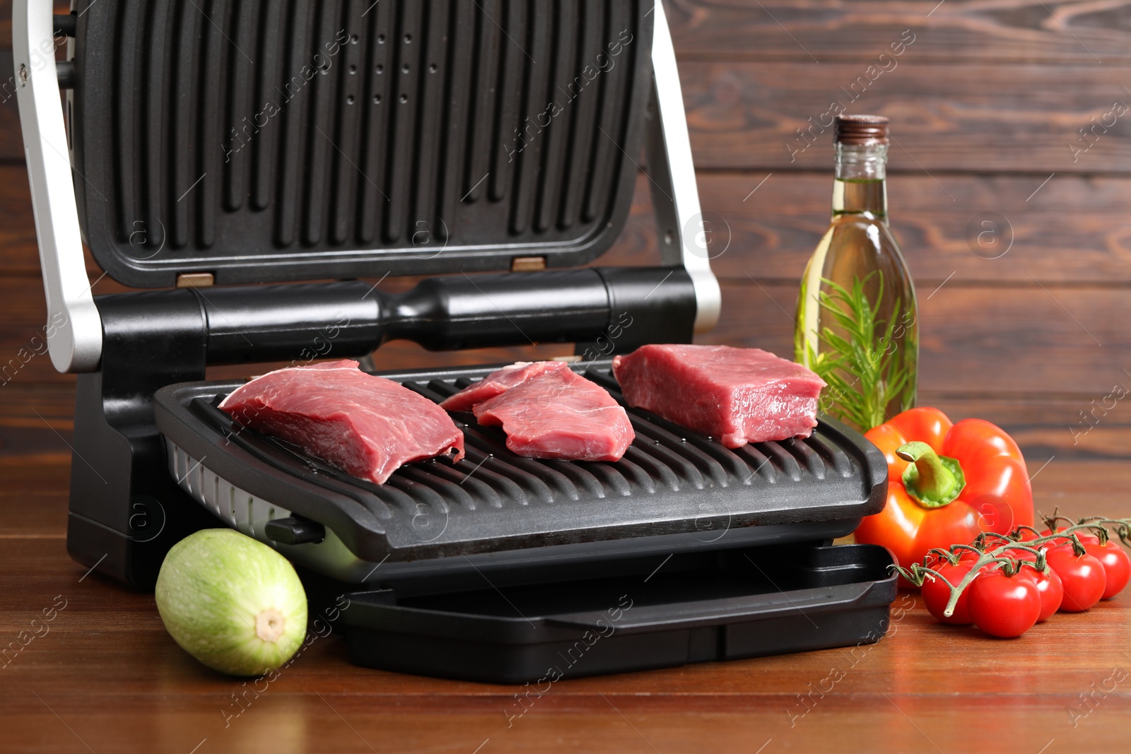 Photo of Electric grill with raw meat, oil and vegetables on wooden table