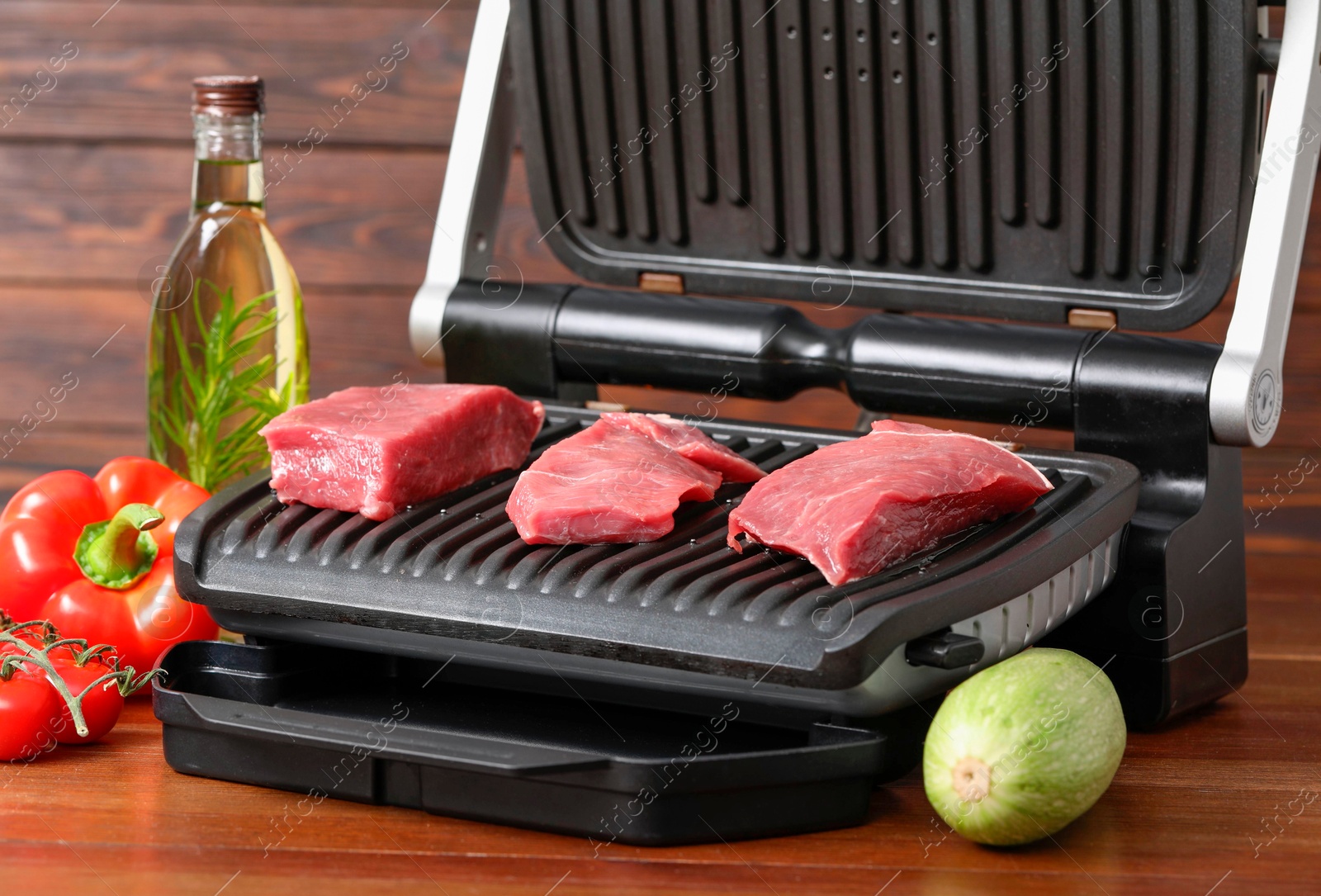 Photo of Electric grill with raw meat, oil and vegetables on wooden table