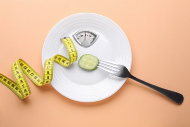 Image of White plate with analog weighing scale display, cucumber slice, fork and measuring tape on beige background, top view