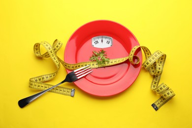 Red plate with analog weighing scale display, arugula, fork and measuring tape on yellow background, top view