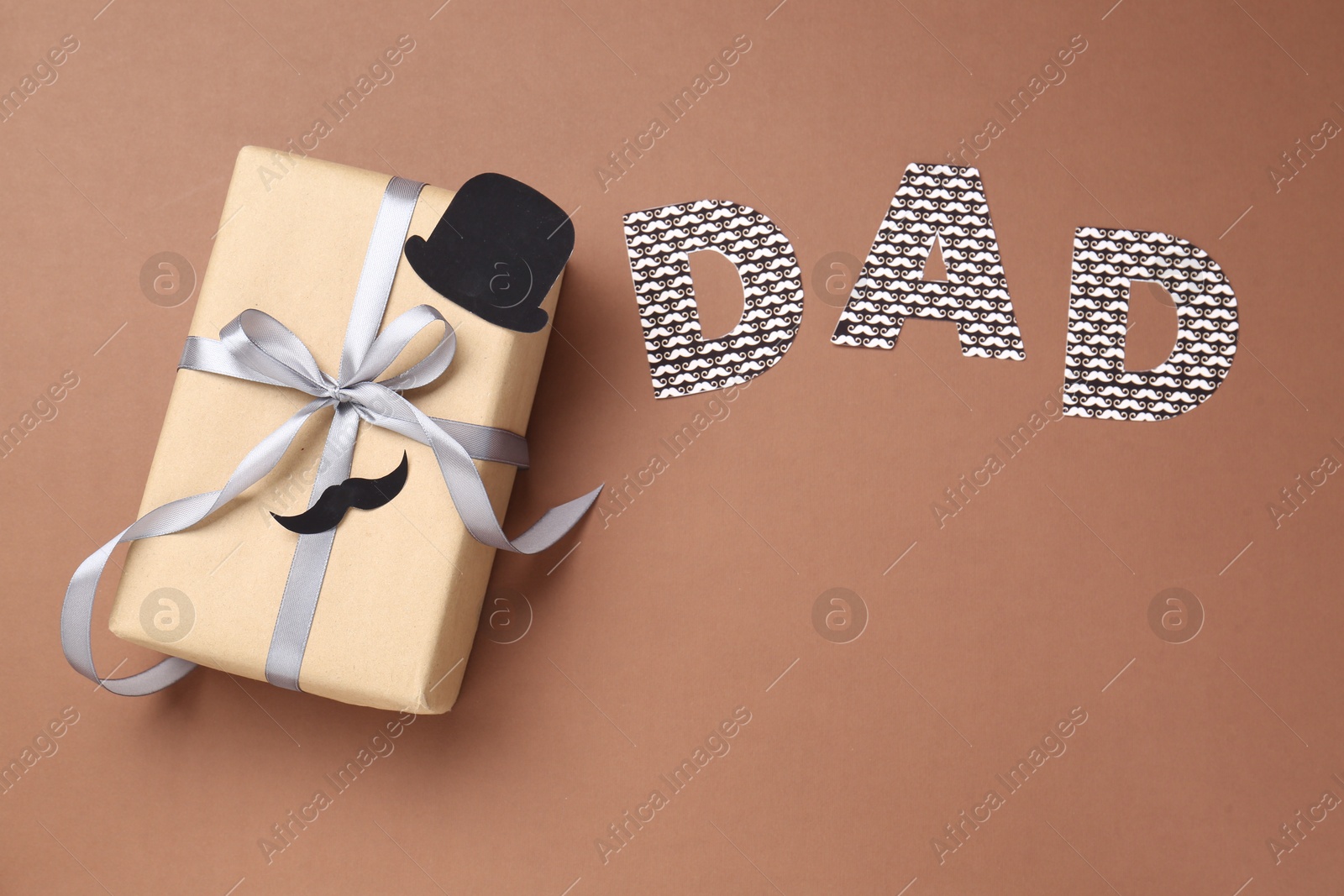 Photo of Happy Father's Day. Gift box with paper hat, moustache and word Dad on brown background, top view