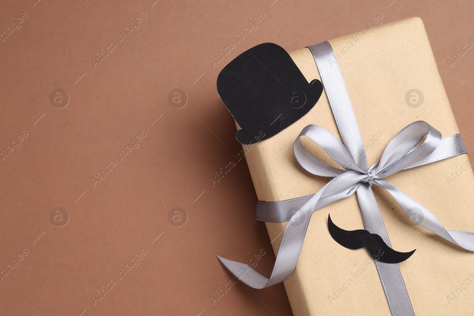 Photo of Happy Father's Day. Gift box with paper hat and moustache on brown background, top view