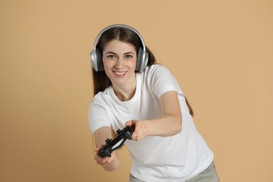 Happy woman in headphones playing video games with controller on beige background