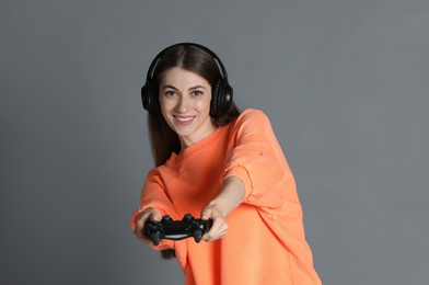 Happy woman in headphones playing video games with controller on gray background