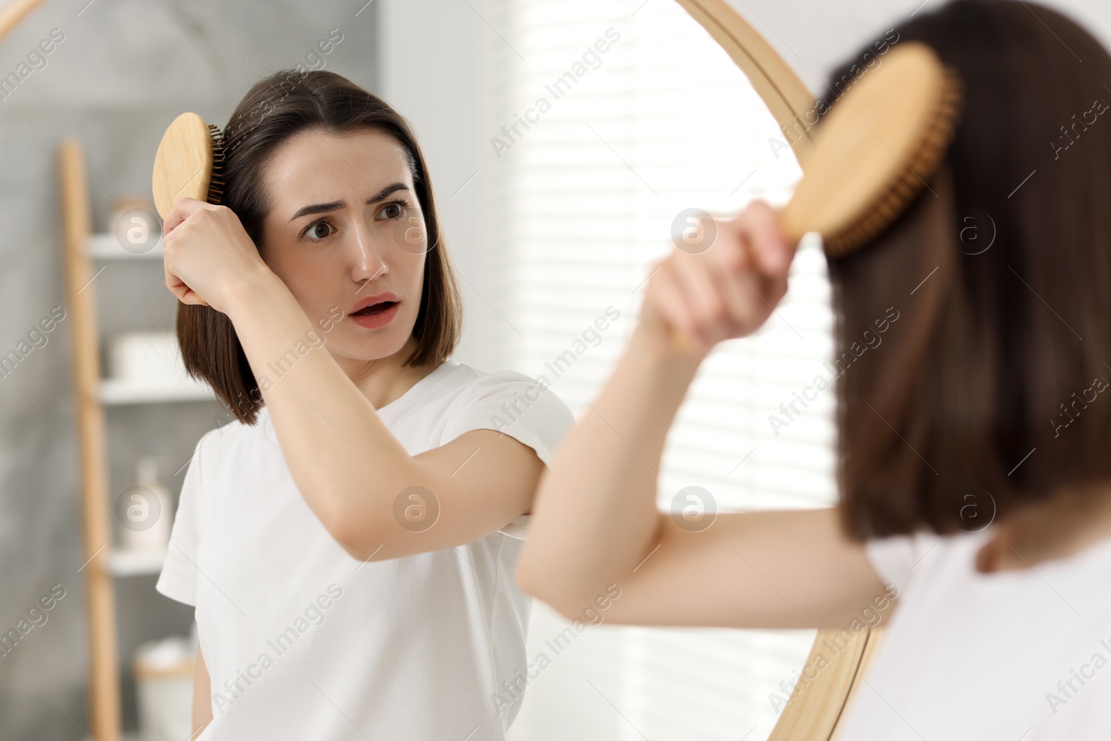 Photo of Emotional woman brushing her hair near mirror indoors. Alopecia problem