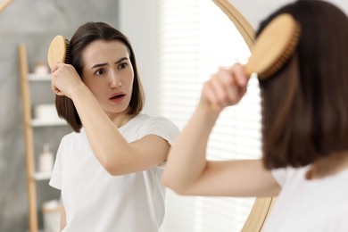 Photo of Emotional woman brushing her hair near mirror indoors. Alopecia problem