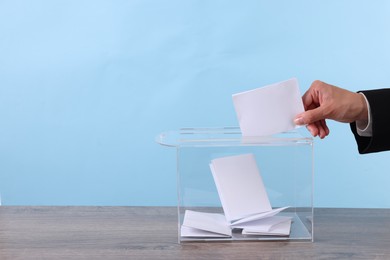 Photo of Woman putting her vote into ballot box at wooden table against light blue background, closeup. Space for text