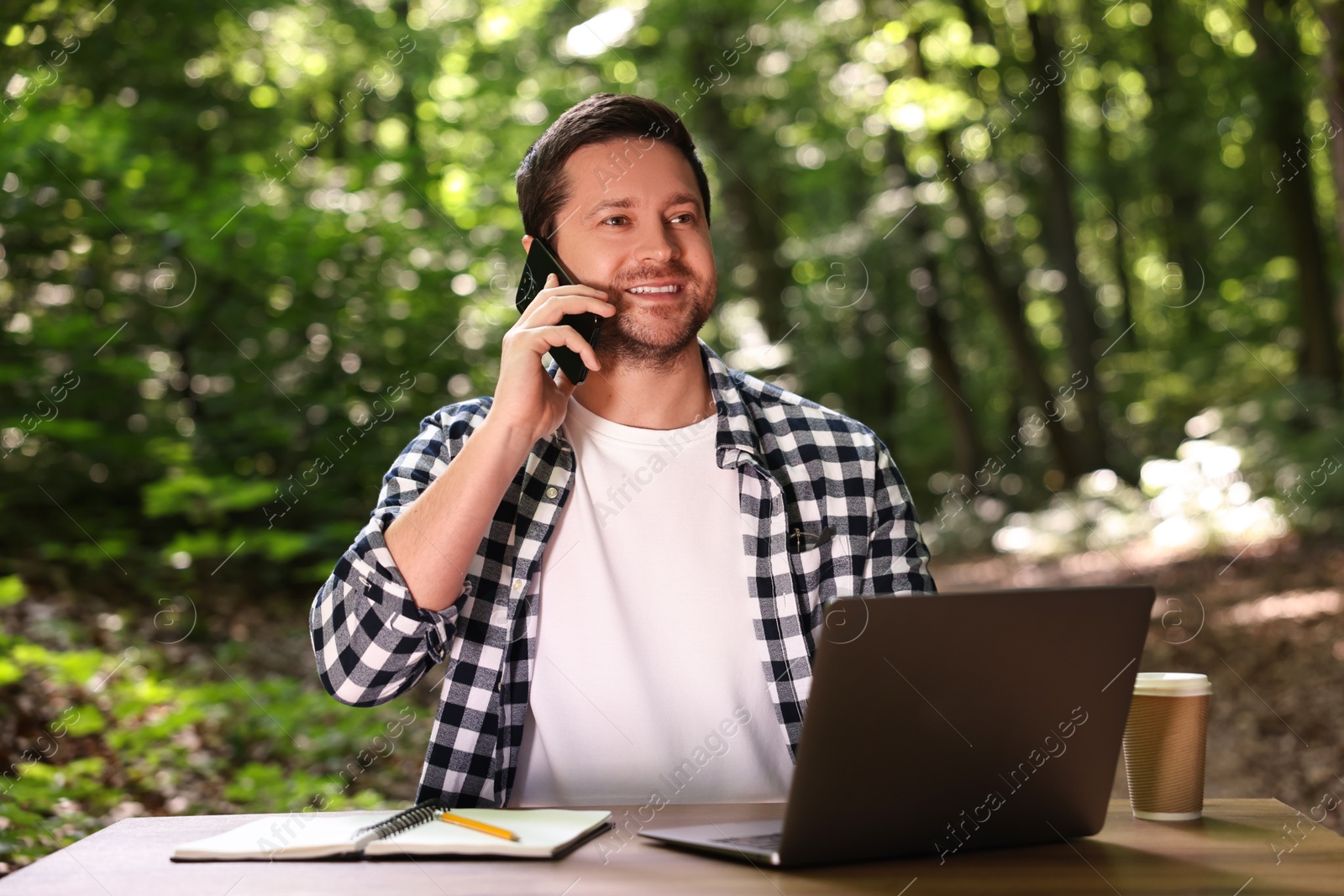 Photo of Smiling freelancer talking on smartphone at table with laptop outdoors. Remote job