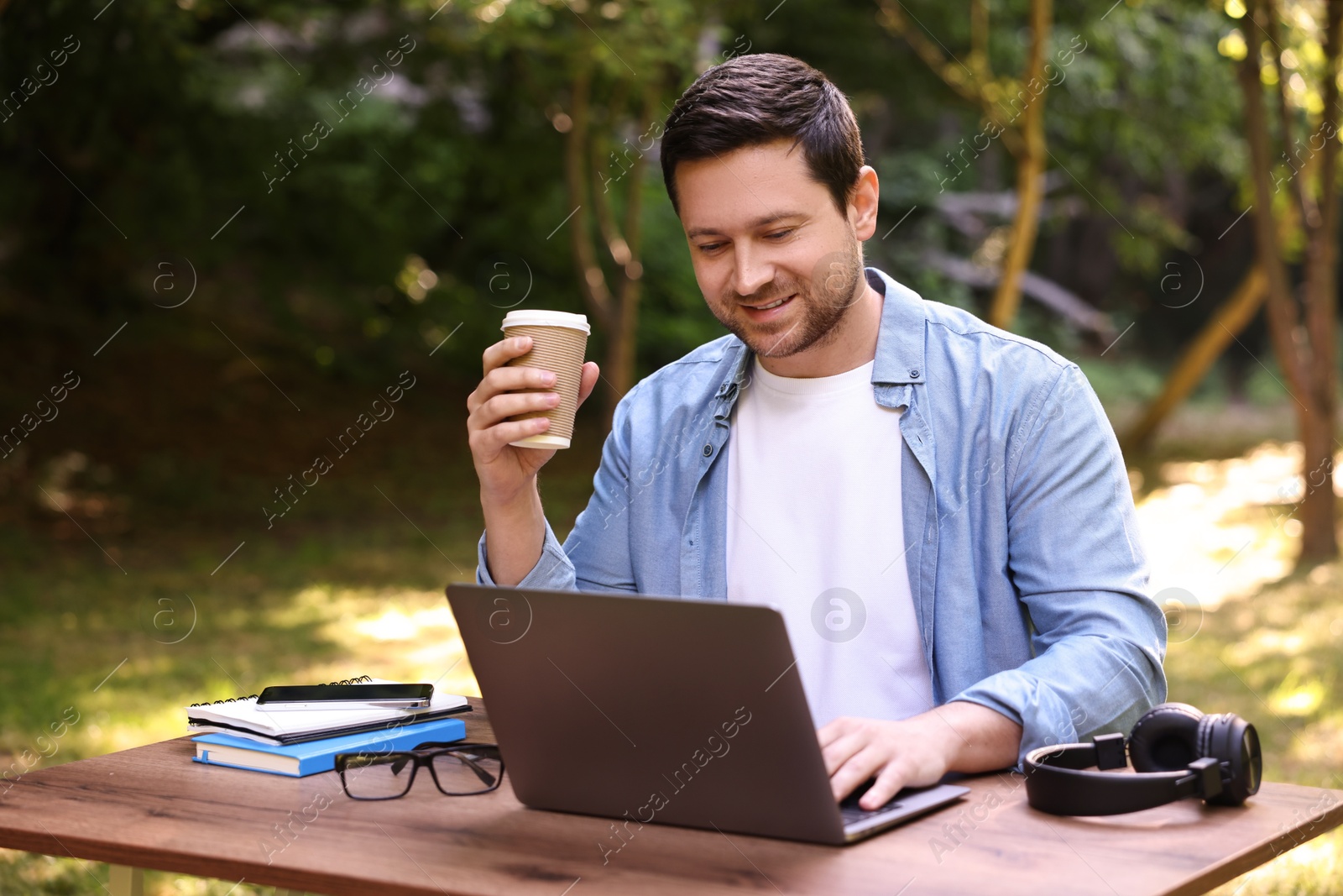 Photo of Smiling freelancer drinking coffee at table with laptop outdoors. Remote job