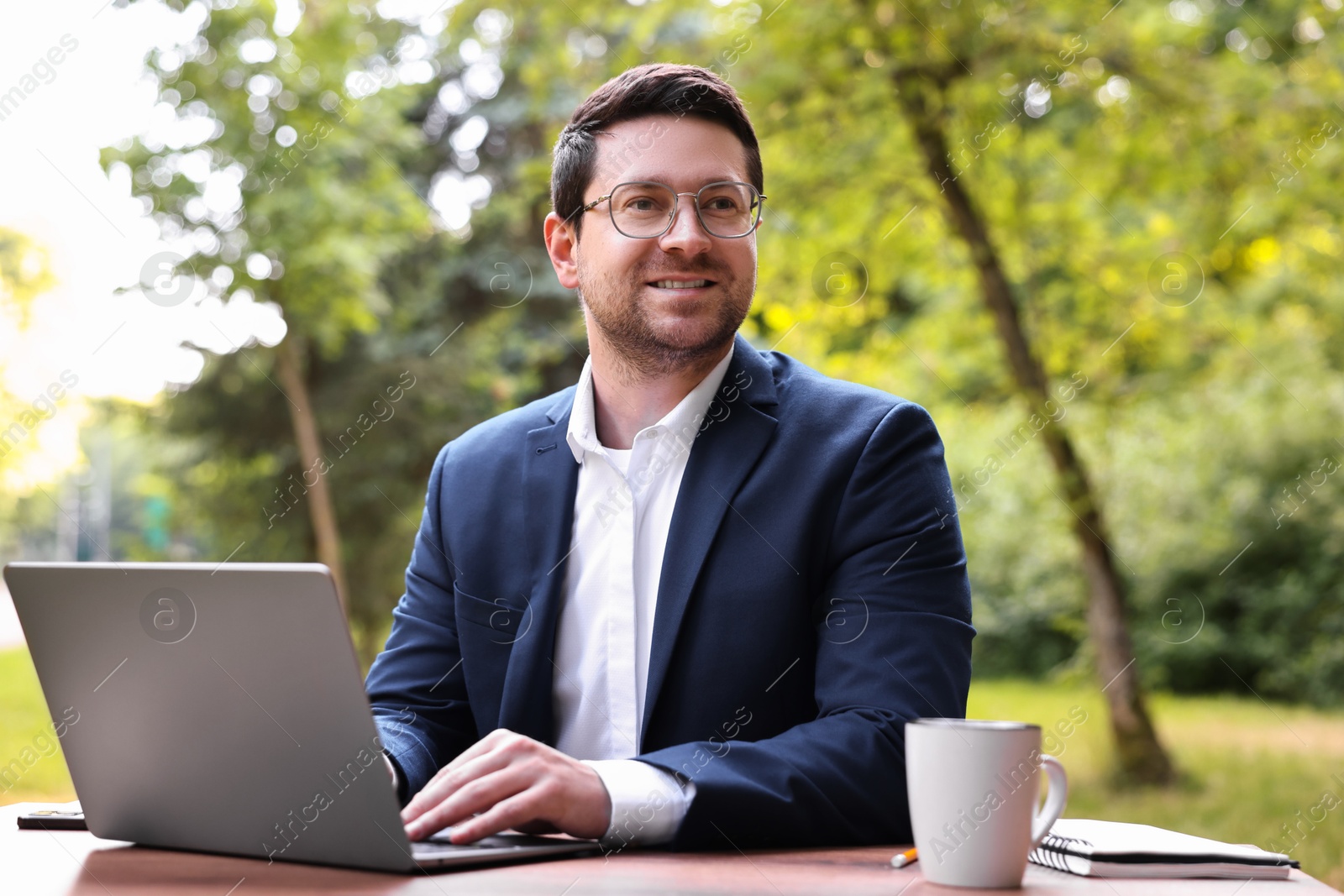 Photo of Smiling businessman working with laptop at table outdoors. Remote job