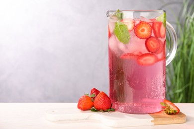 Photo of Freshly made strawberry lemonade with mint in jug and glass on white wooden table against grey background, space for text