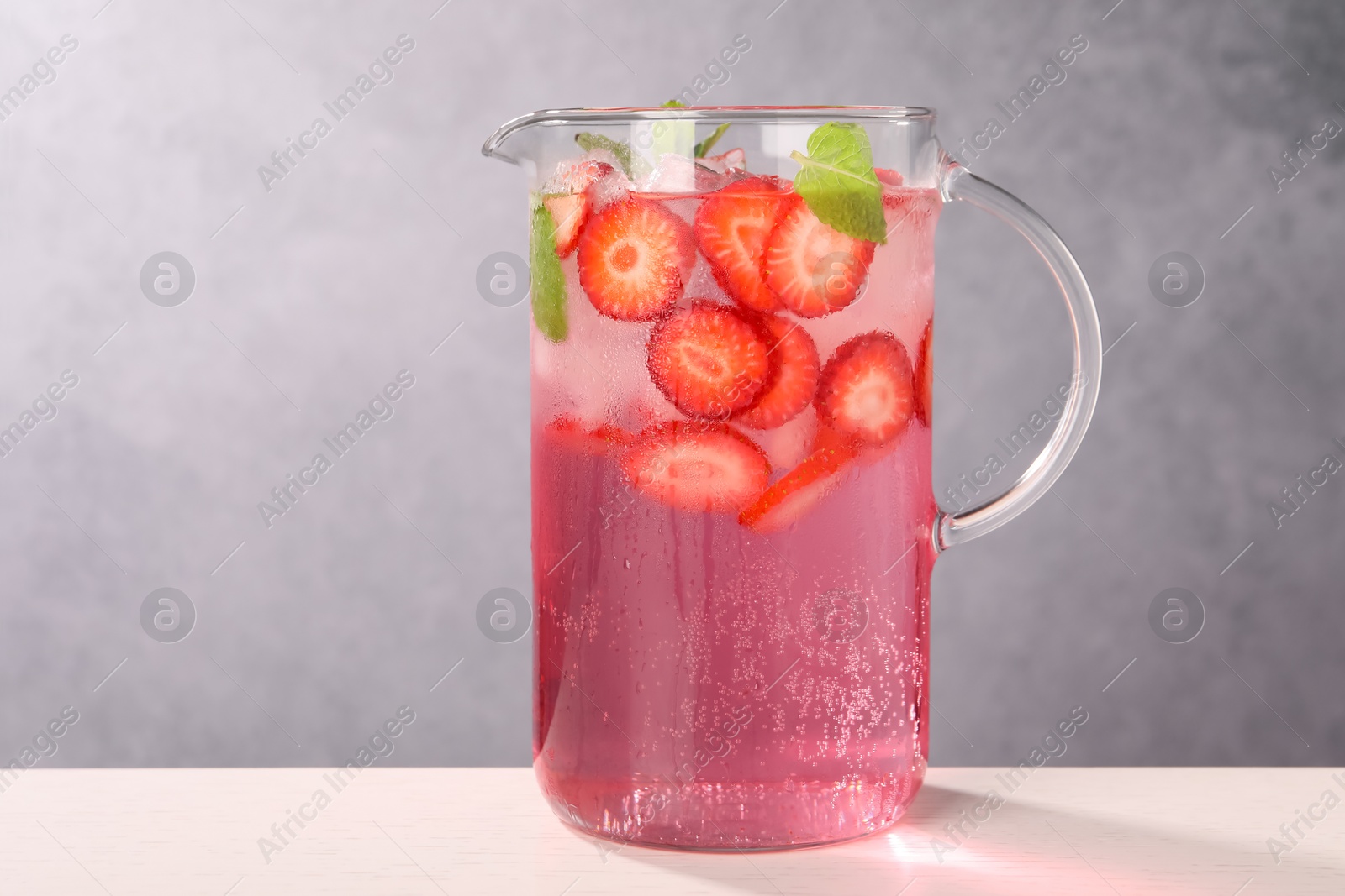 Photo of Freshly made strawberry lemonade with mint in jug and glass on white wooden table against grey background