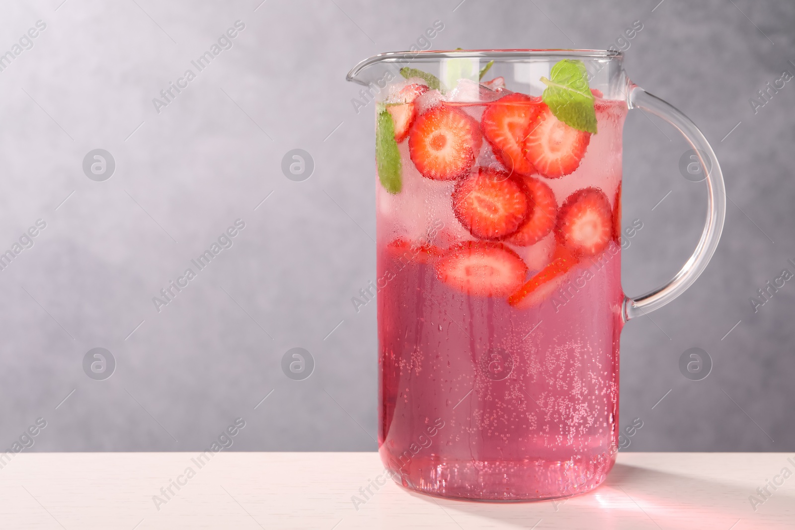 Photo of Freshly made strawberry lemonade with mint in jug and glass on white wooden table against grey background, space for text