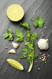Fresh coriander leaves, dried seeds, garlic and lime on black textured table, flat lay