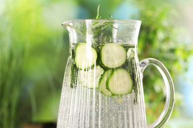 Refreshing cucumber water with rosemary in jug against blurred green background, closeup