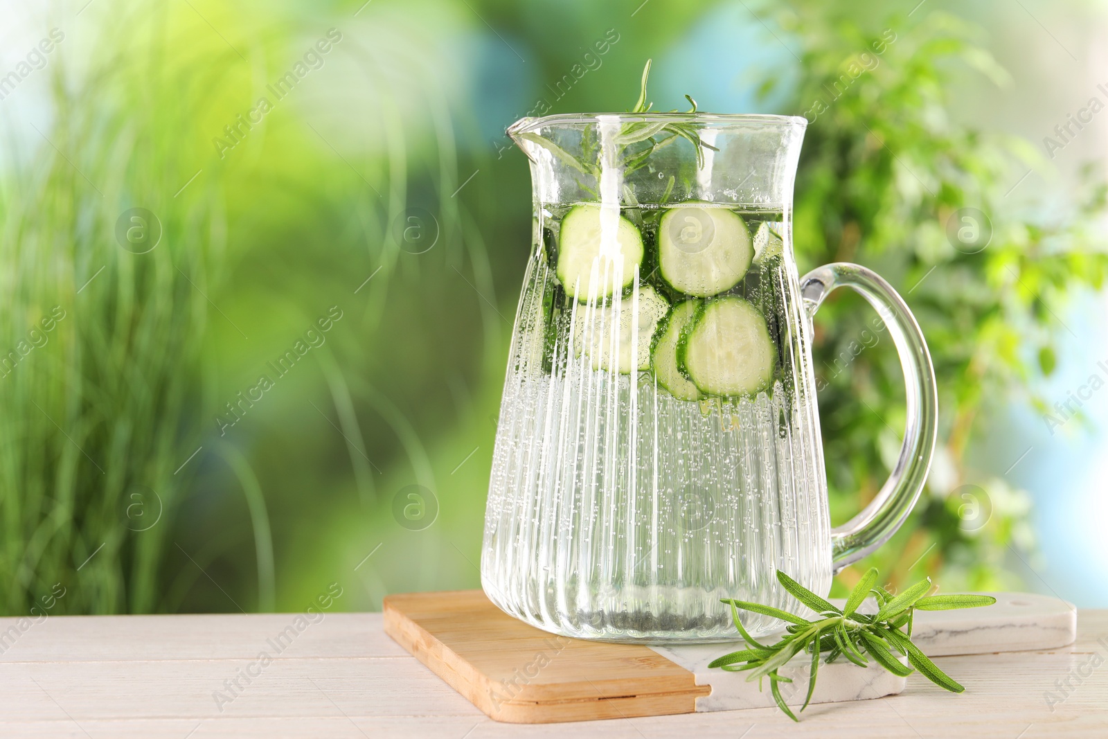 Photo of Refreshing cucumber water with rosemary in jug on light wooden table against blurred green background. Space for text