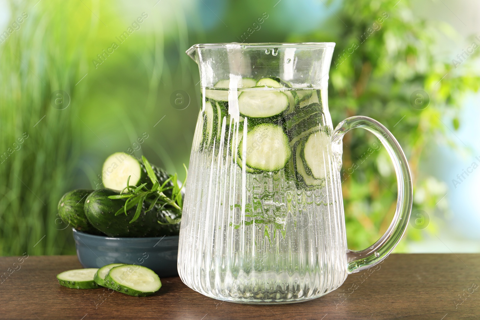 Photo of Refreshing cucumber water with rosemary in jug and vegetable on wooden table against blurred green background