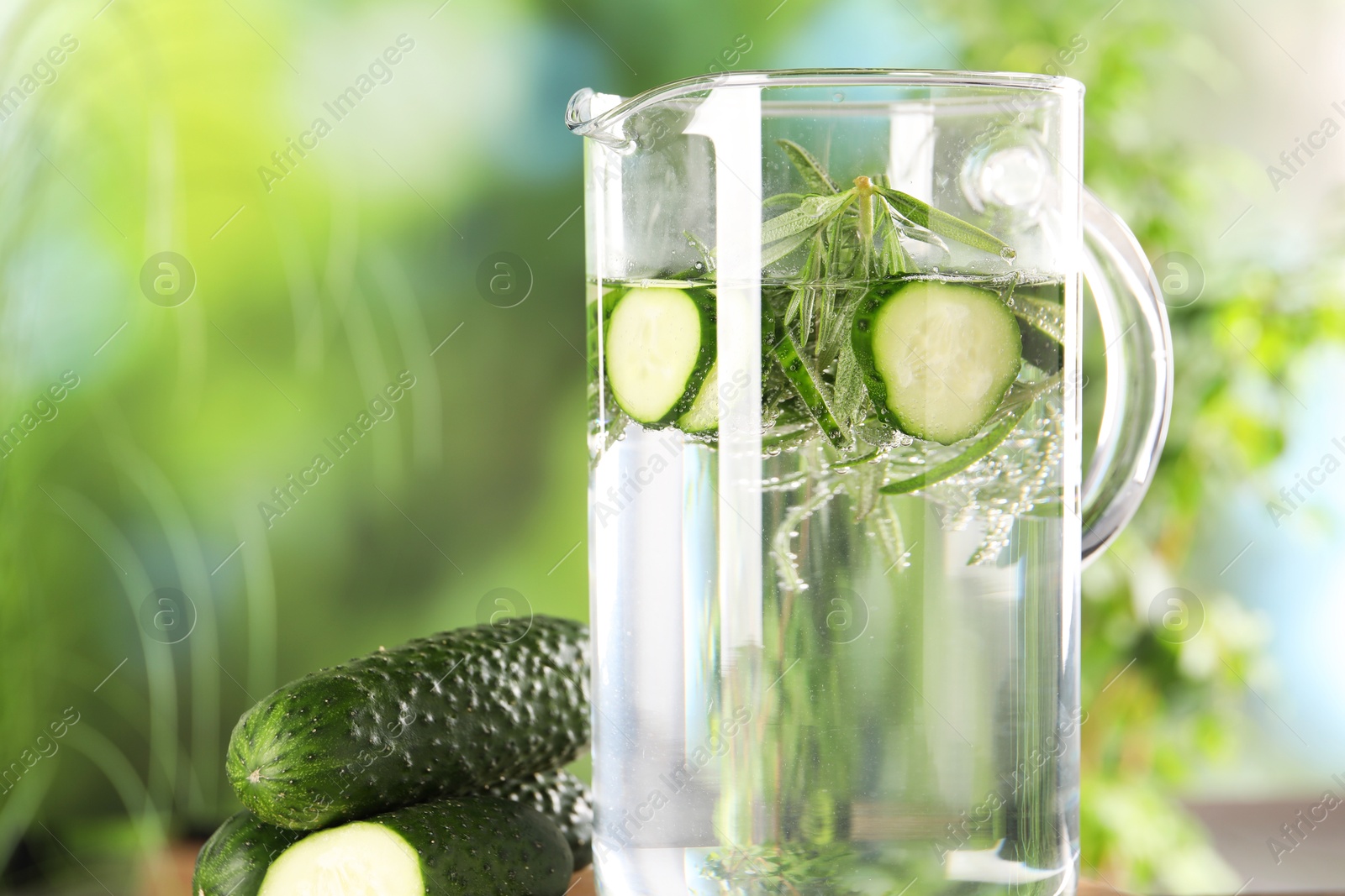 Photo of Refreshing cucumber water with rosemary in jug and vegetable against blurred green background, closeup