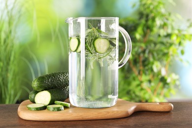 Refreshing cucumber water with rosemary in jug and vegetable on wooden table against blurred green background