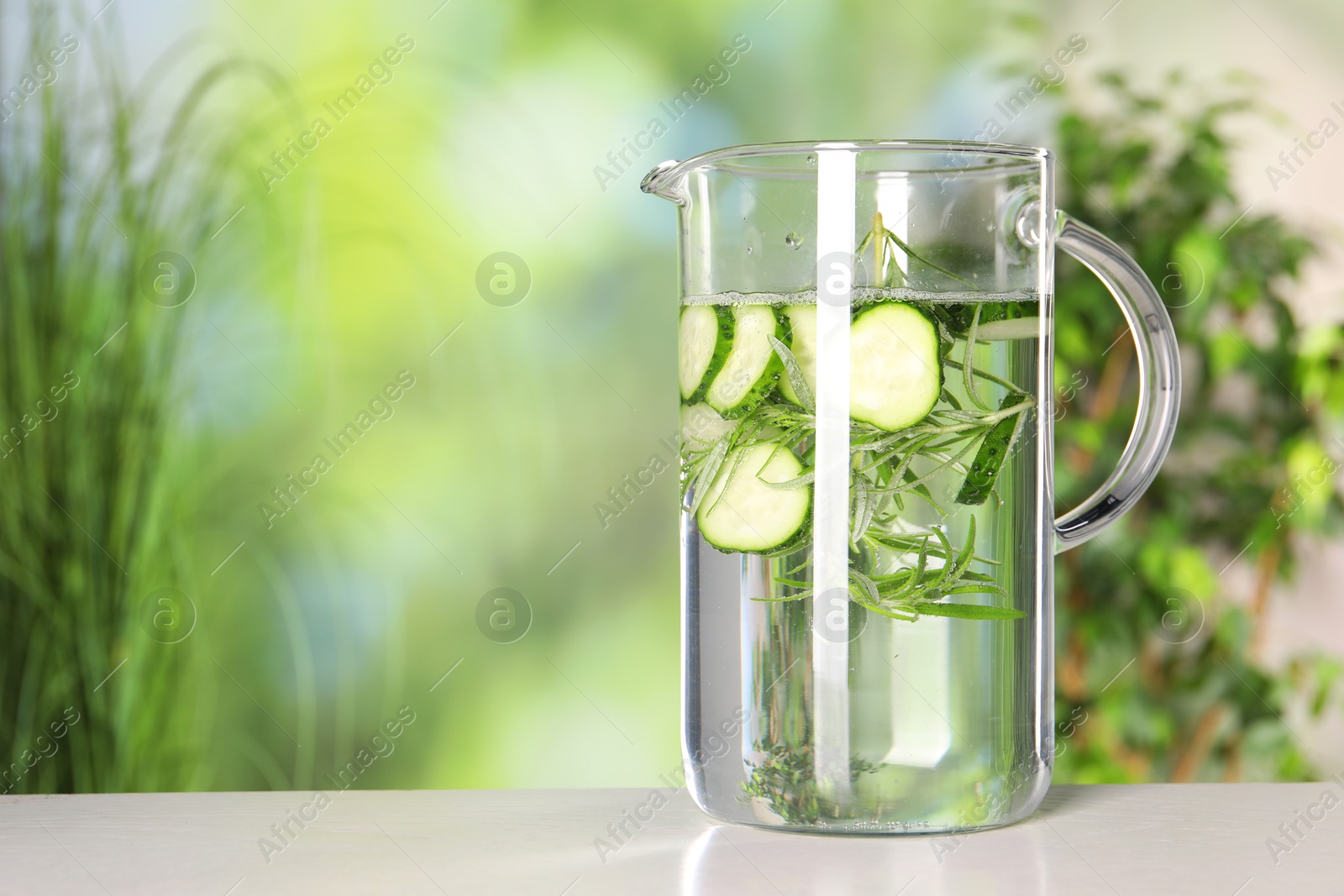 Photo of Refreshing cucumber water with rosemary in jug on light table against blurred green background. Space for text