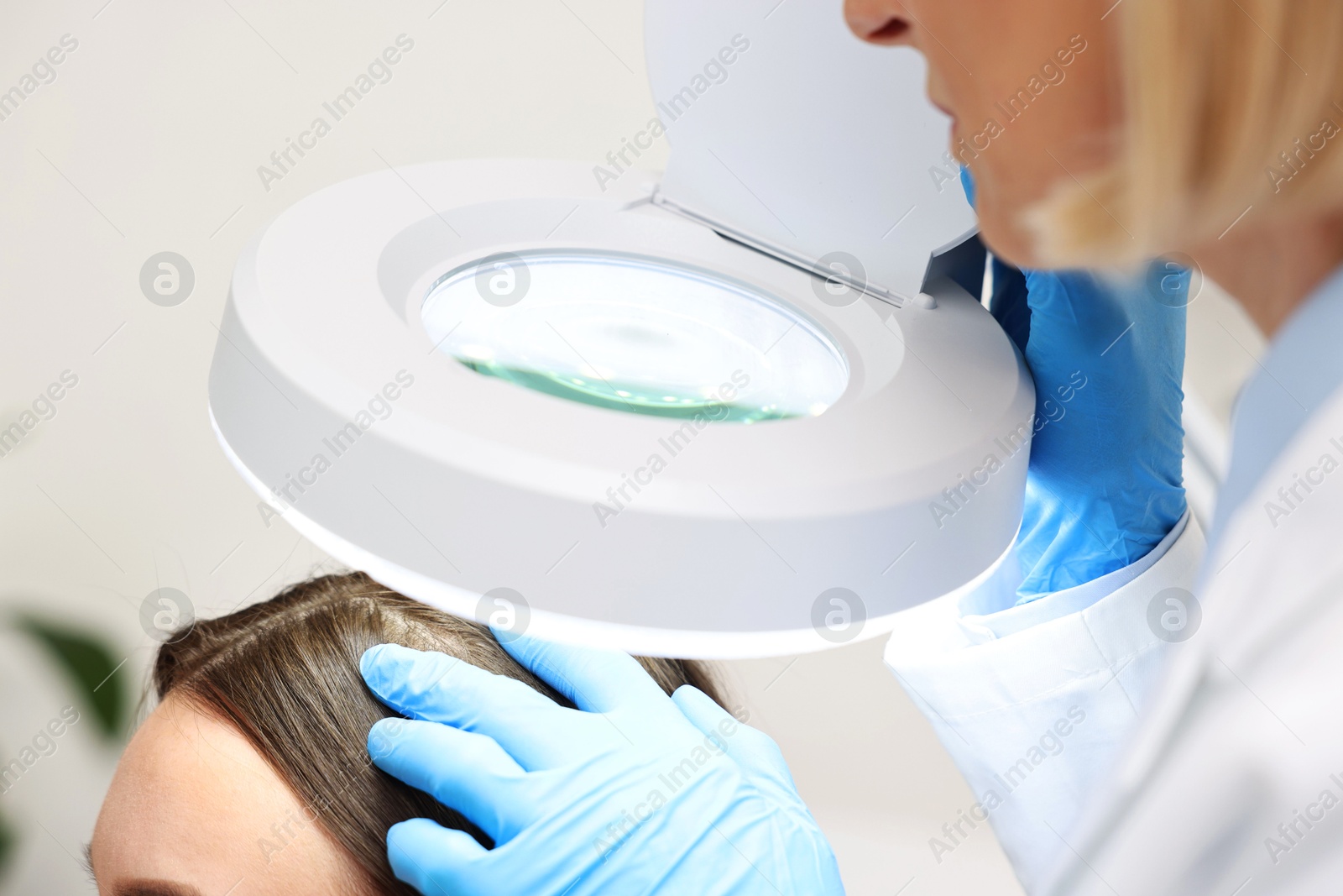 Photo of Trichologist examining patient`s hair under lamp in clinic, closeup