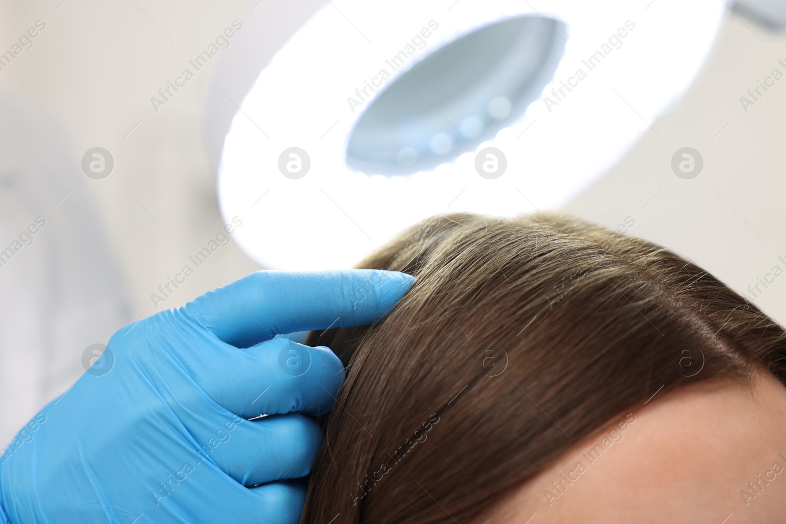 Photo of Trichologist examining patient`s hair under lamp in clinic, closeup
