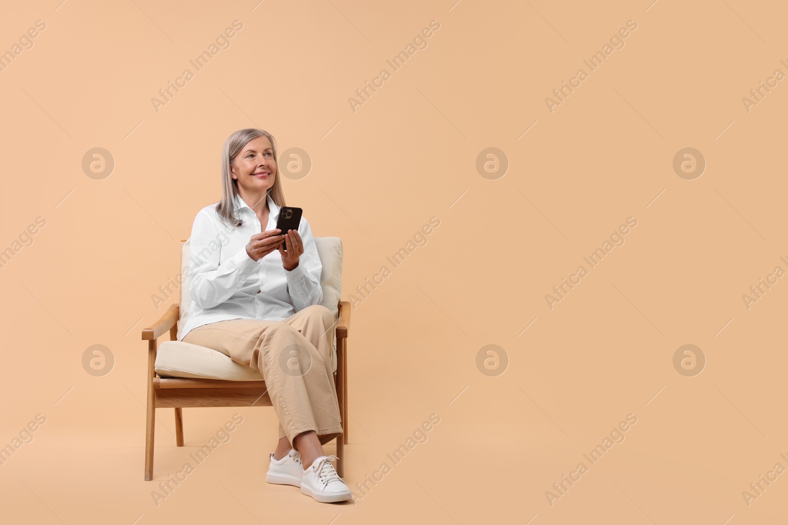 Photo of Senior woman with phone on armchair against beige background, space for text