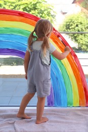Photo of Little girl drawing rainbow on window indoors, back view