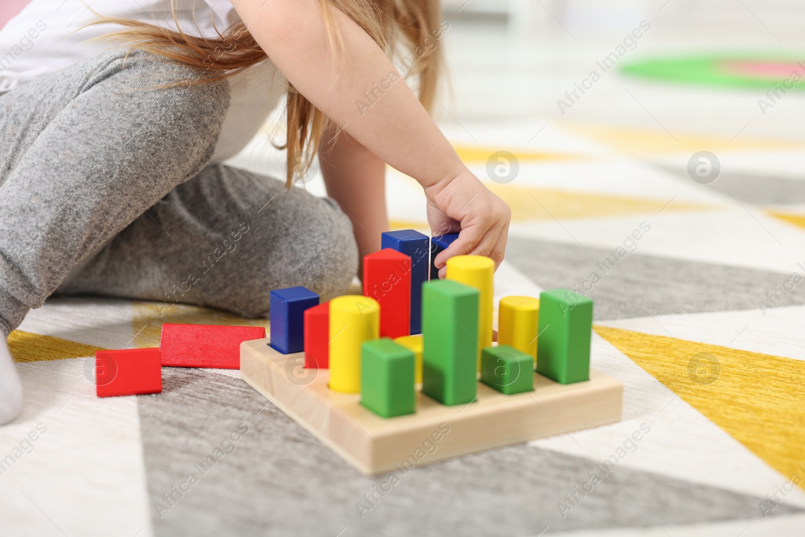 Photo of Little girl playing with set of wooden geometric figures on carpet, closeup. Kindergarten activities for motor skills development