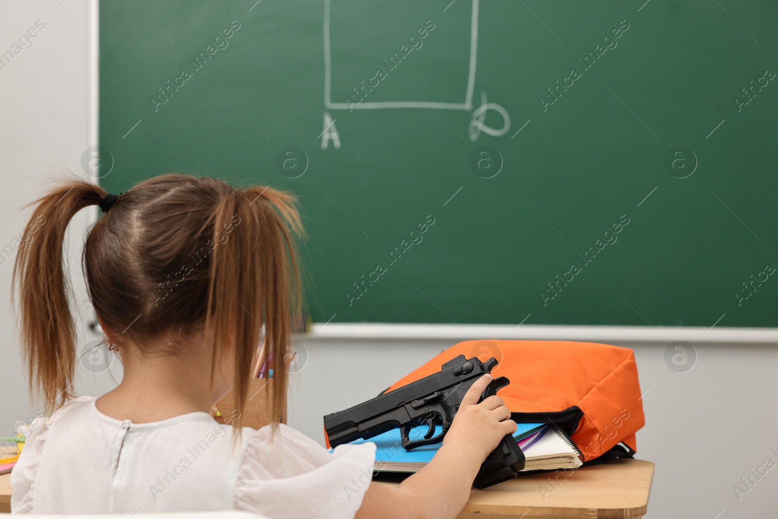 Photo of Child with gun and school stationery at desk in classroom