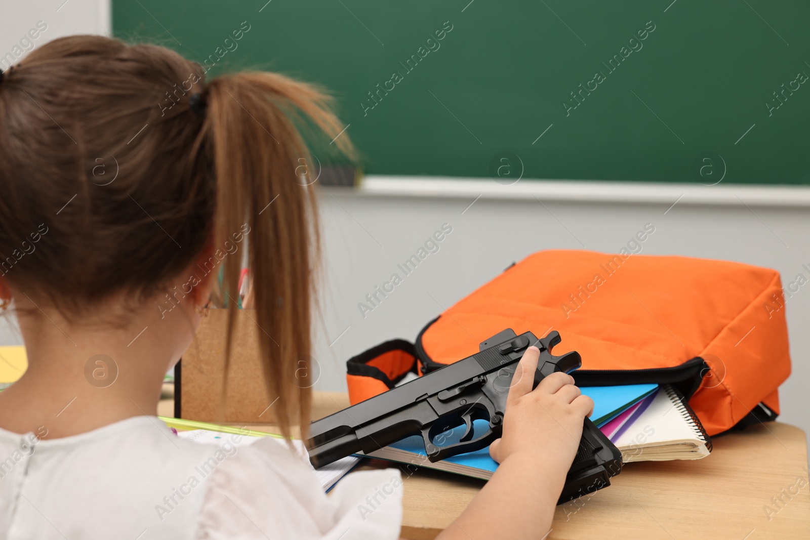 Photo of Child with gun and school stationery at desk in classroom