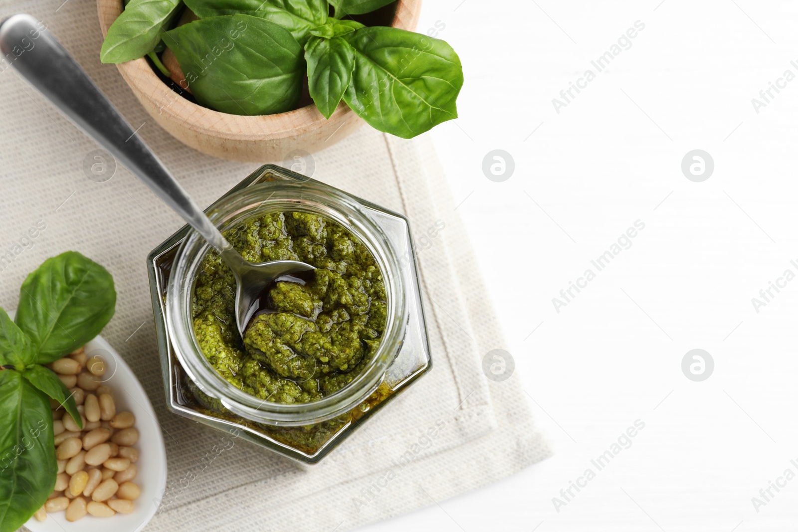 Photo of Tasty pesto sauce in jar, spoon, pine nuts and basil on white table, top view. Space for text